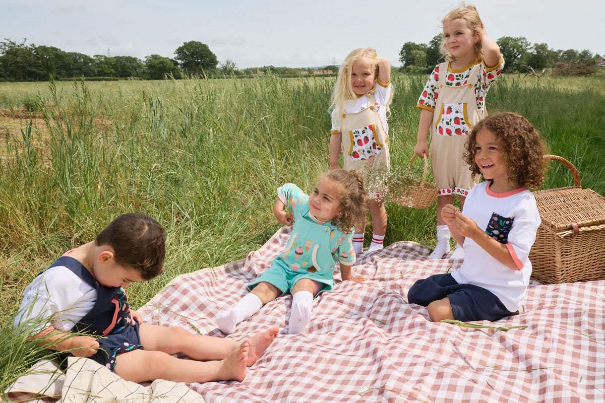 A group of 5 children gathered on a picnic blanket in the middle of a green field. They are wearing a selection of Playtime Prints outfits including strawberry, fish and ice cream prints.