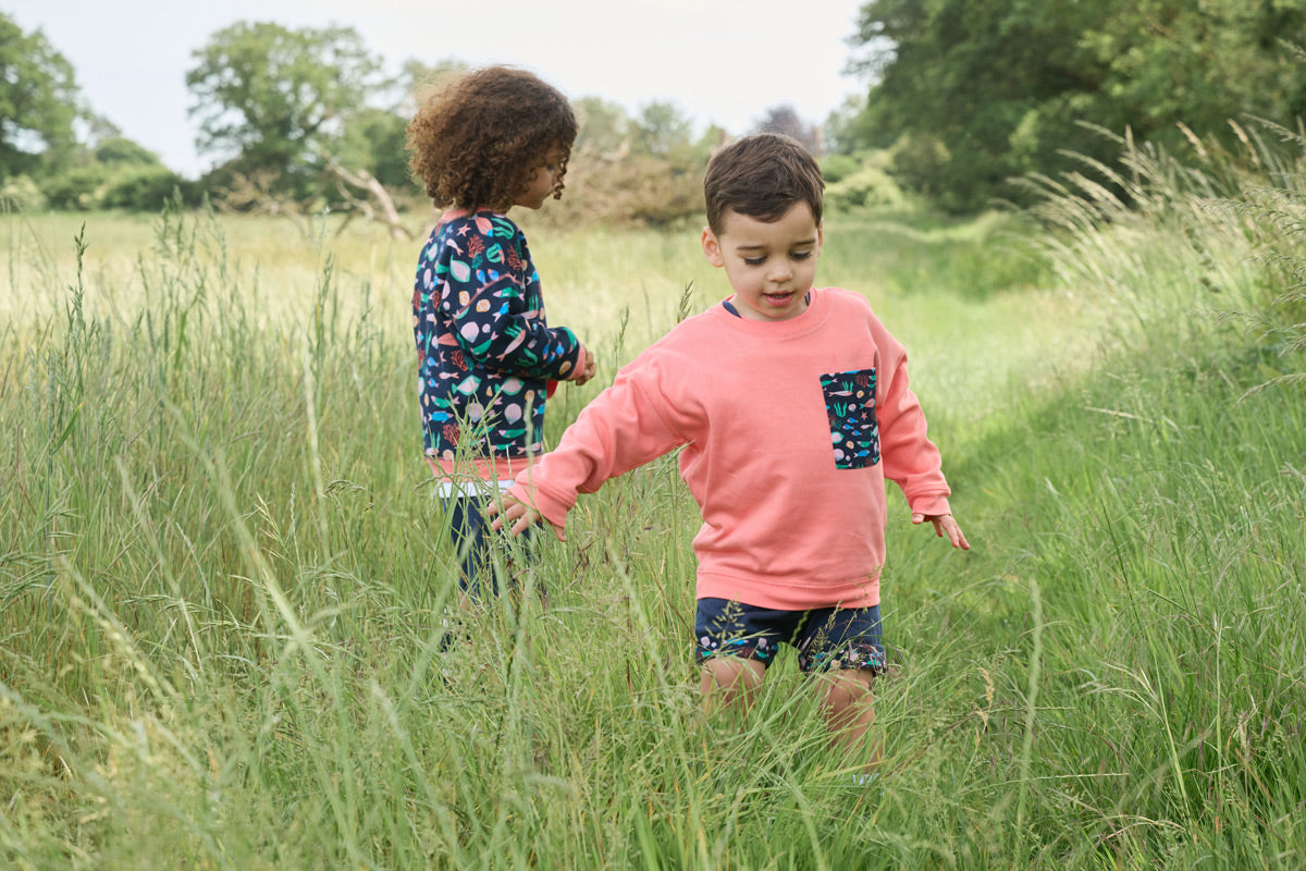 Two boys playing in a field - one wearing a sweatshirt featuring an all over vibrant navy fish print with contrast coral neck and sleeve trim and the other a coral sweatshirt with an oversized vibrant navy fish print chest pocket.