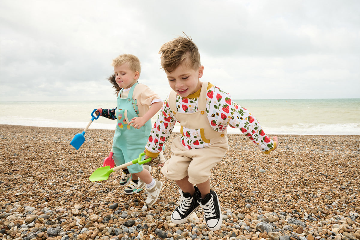 Three boys jumping up into the air on a pebble beach in front of the sea. They are wearing bright and vibrant Playful Prints outfits featuring ice cream, strawberry and fish prints.