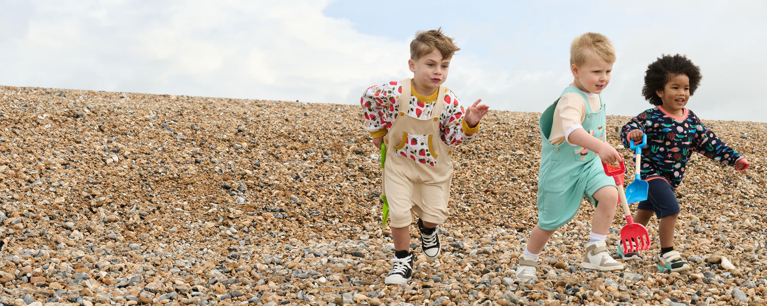 Three young boys running down a pebble beach with shovels and spades. The first boy is wearing a strawberry print outfit, the second is an ice cream print set and the third a fish print.