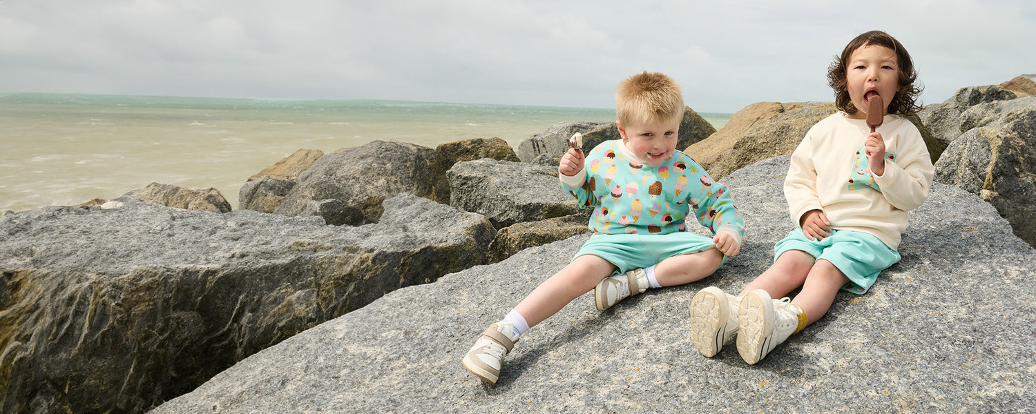 Two boys sitting eating ice cream on some large rocks by the sea. They are wearing vibrant turquoise clothes featuring an ice cream print.