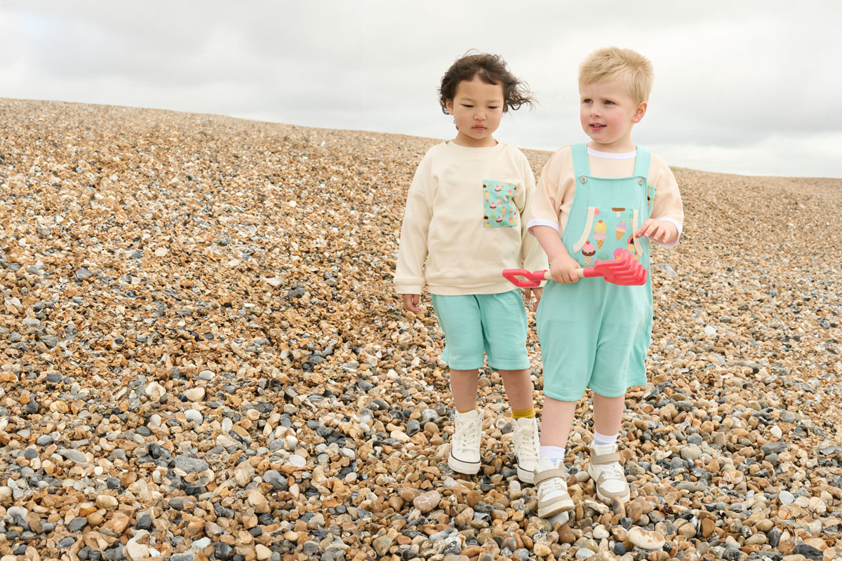 Two young boys playing on a pebble beach wearing matching outfits featuring a turquoise ice cream print.