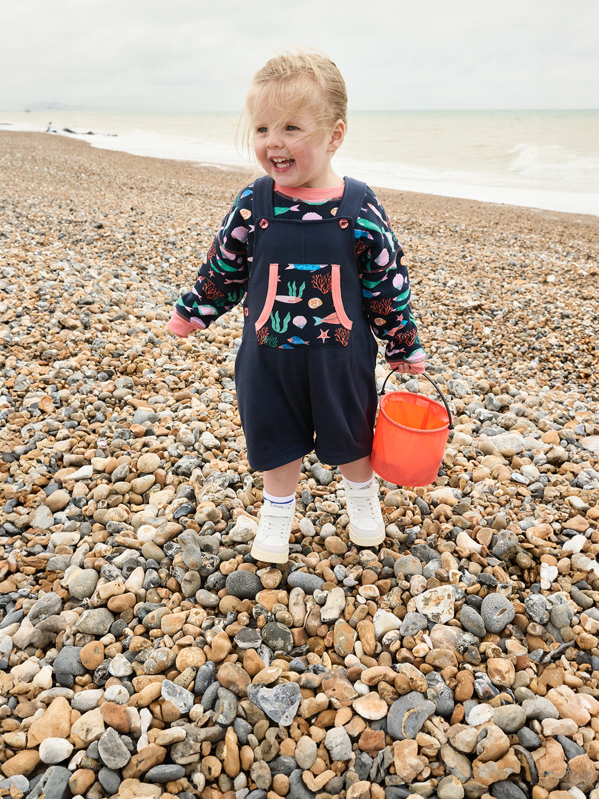 Little girl on a pebble beach in front of the sea holding a bucket filled with pebbles. She is smiling and wearing a vibrant matching sweatshirt and dungarees set in the Playtime Prints fish design.