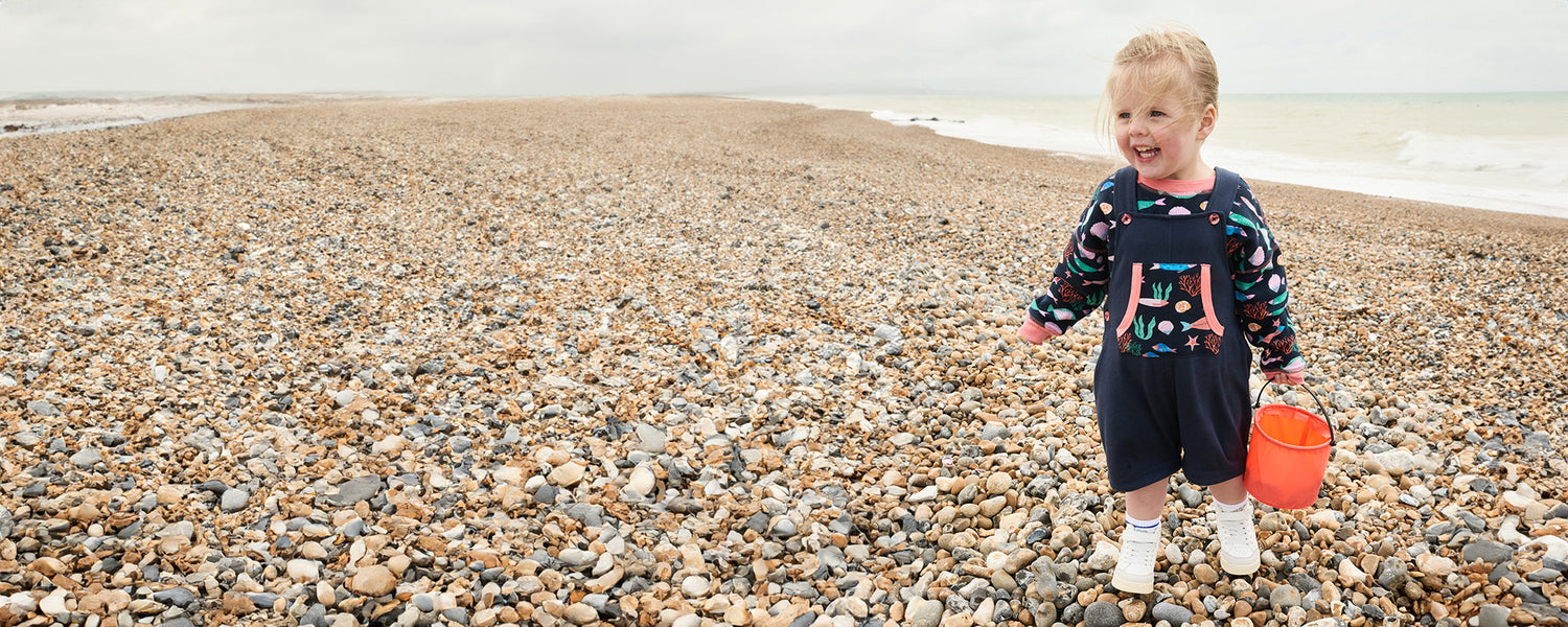 Little girl on a pebble beach in front of the sea holding a bucket filled with pebbles. She is smiling and wearing a vibrant matching sweatshirt and dungarees set in the Playtime Prints fish design.