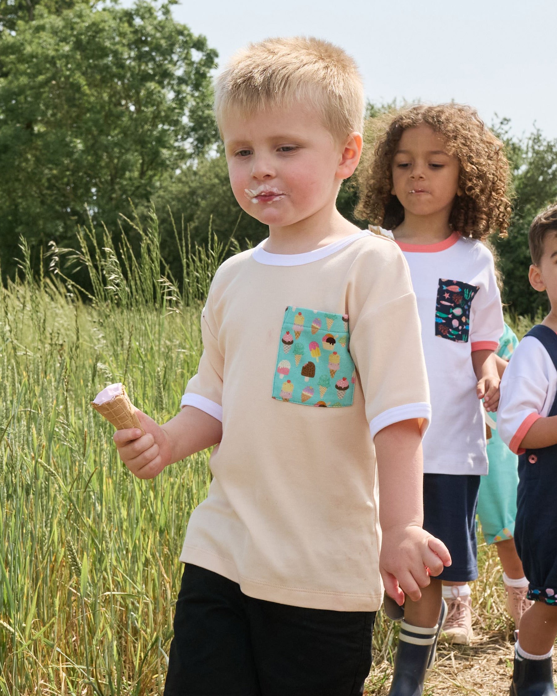 Young boy in a field with his friends eating an ice cream and wearing a vanilla coloured short sleeved t-shirt with bright ice cream print square chest pocket and white neck and sleeve trim over black shorts.