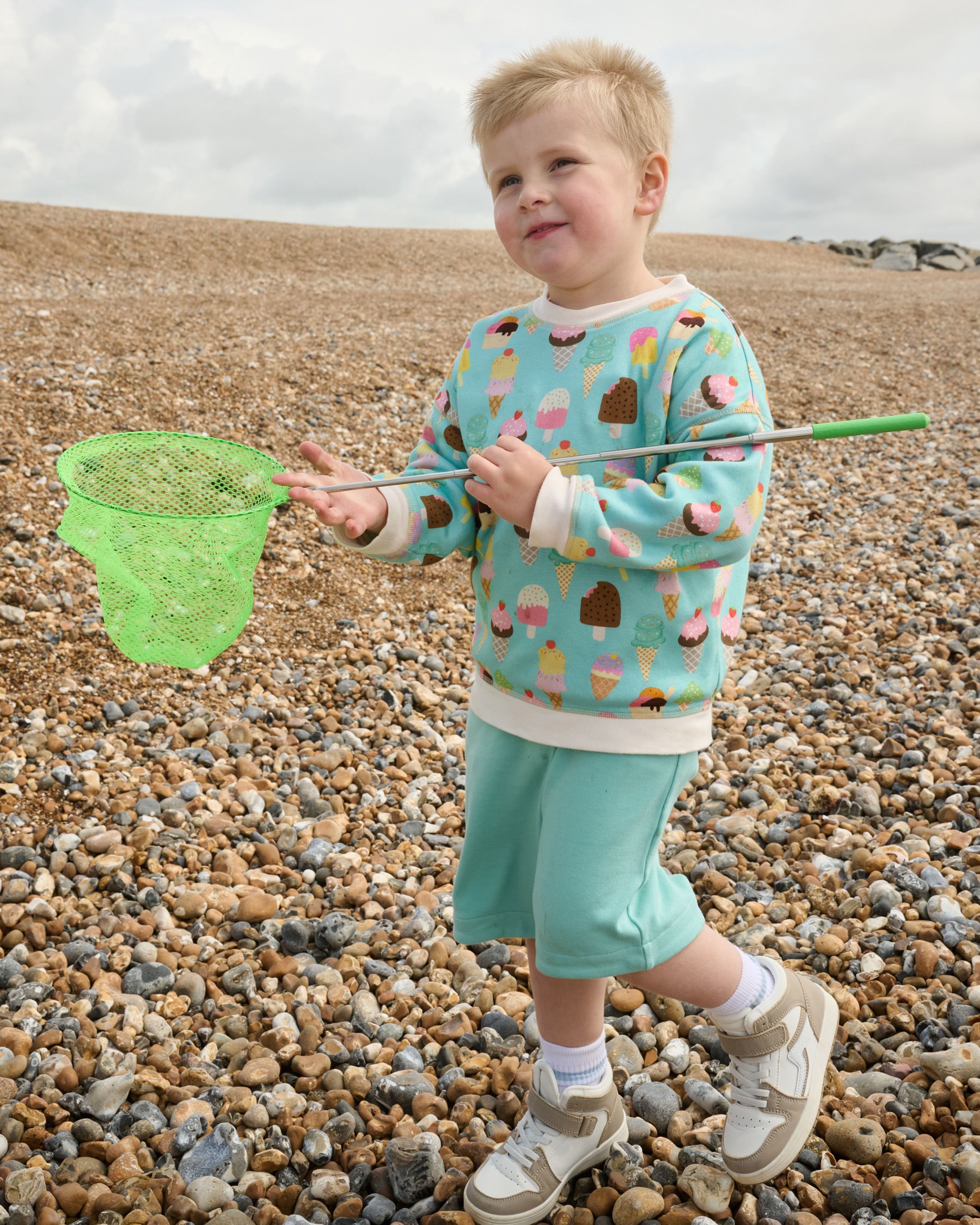 Young smiling boy with a fishing net at the beach wearing a Front of sweatshirt featuring an all over turquoise ice cream print with contrast vanilla neck and sleeve trim over matching turquoise short dungarees.