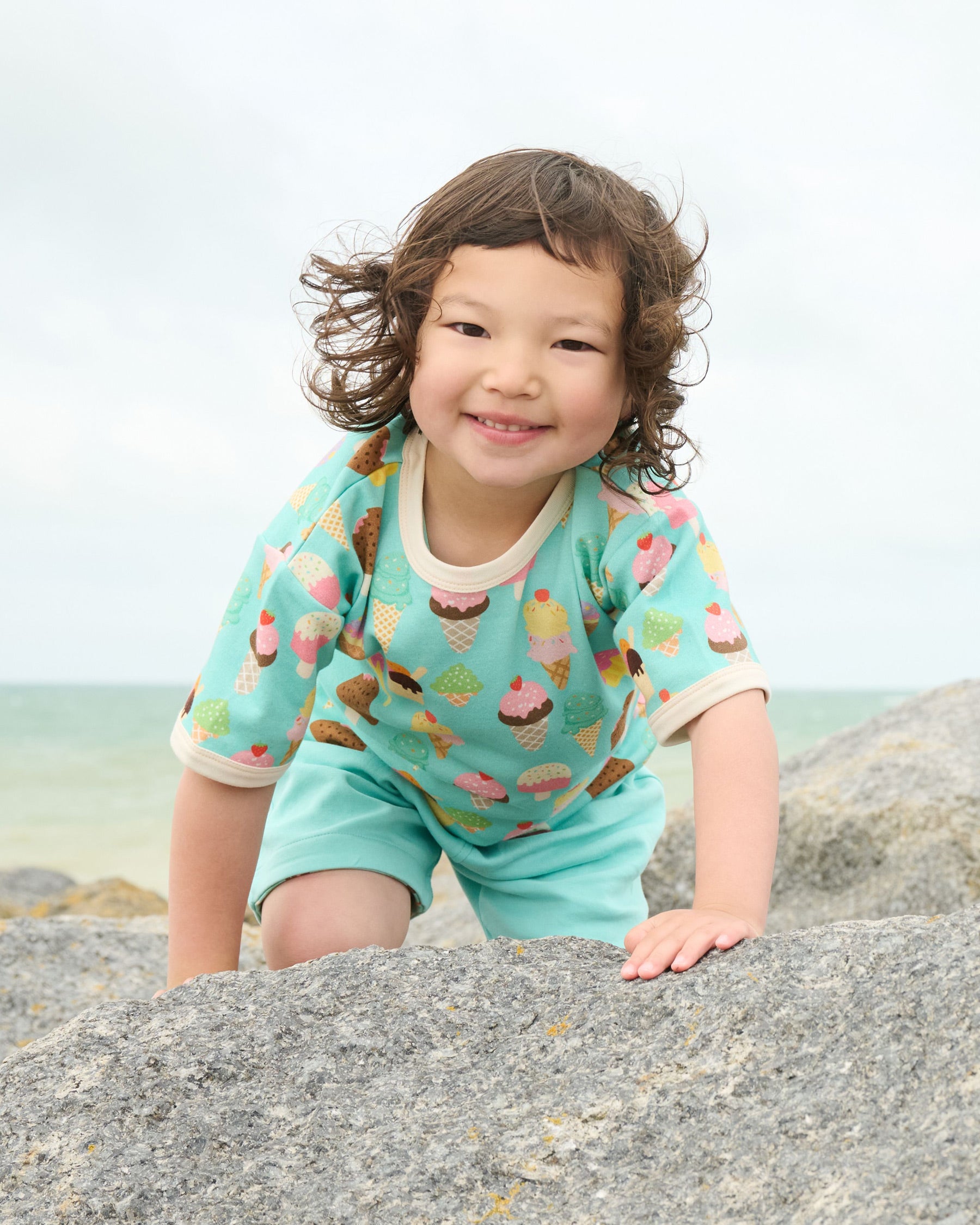 Young boy climbing over rocks at the seaside wearing a short sleeved t-shirt featuring an all over turquoise ice cream print with contrast ecru neck and sleeve trim over a set of matching turquoise short dungarees.