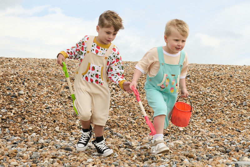 Two young boys running down a pebble beach playing with spades and a bucket. The first boy is wearing a strawberry print outfit and the second is wearing an ice cream print set.