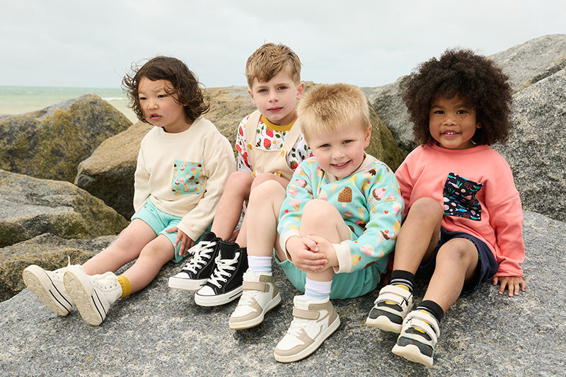 Four boys sitting on large rocks in front of the sea. They are wearing bright and vibrant Playful Prints outfits featuring ice cream, strawberry and fish print sweatshirts.
