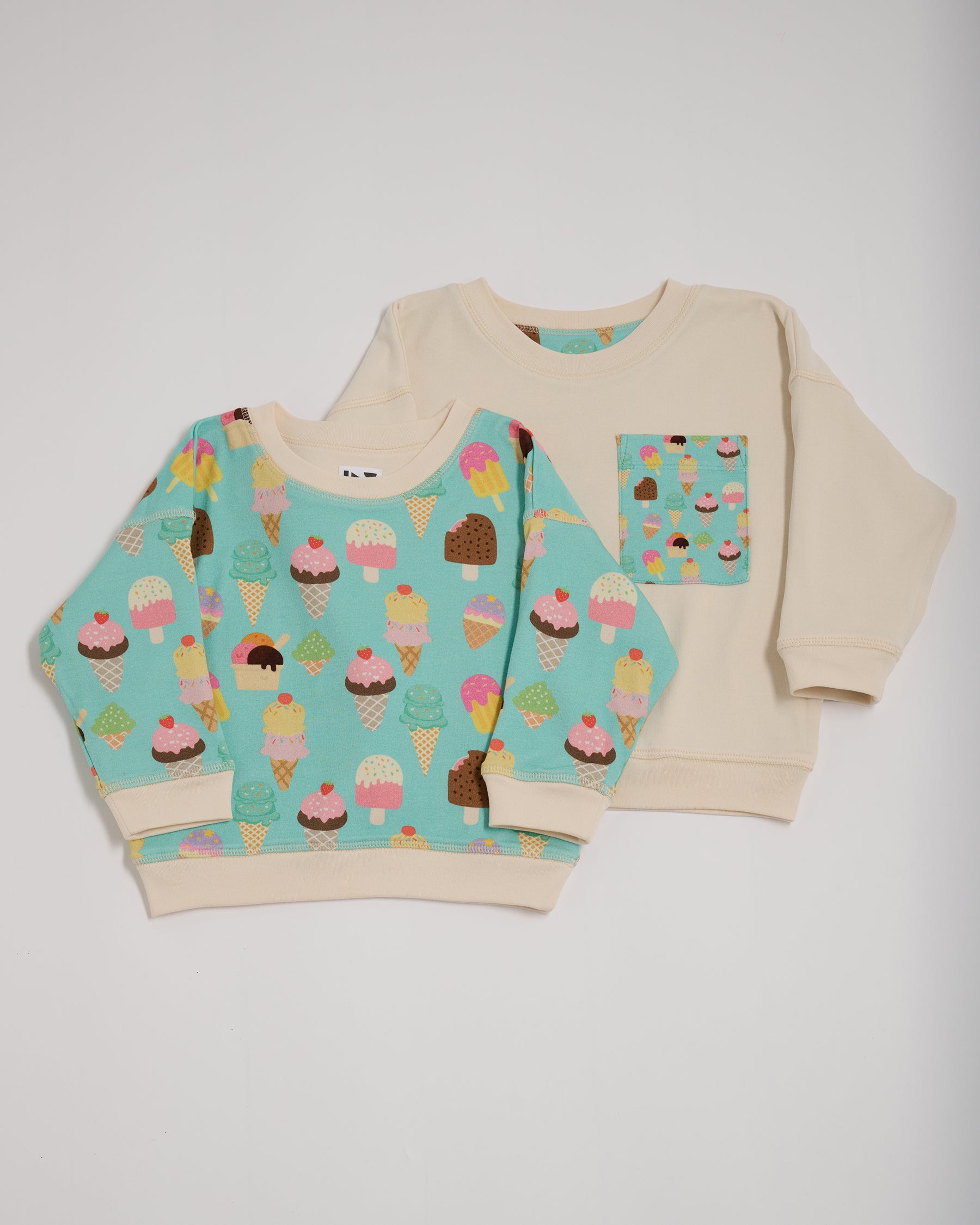 Shoulder to shoulder comparison of both sides of a reversible sweatshirt. One featuring an all over turquoise ice cream print with contrast ecru neck and sleeve trim. The other ecru with an oversized turquoise ice cream print chest pocket.