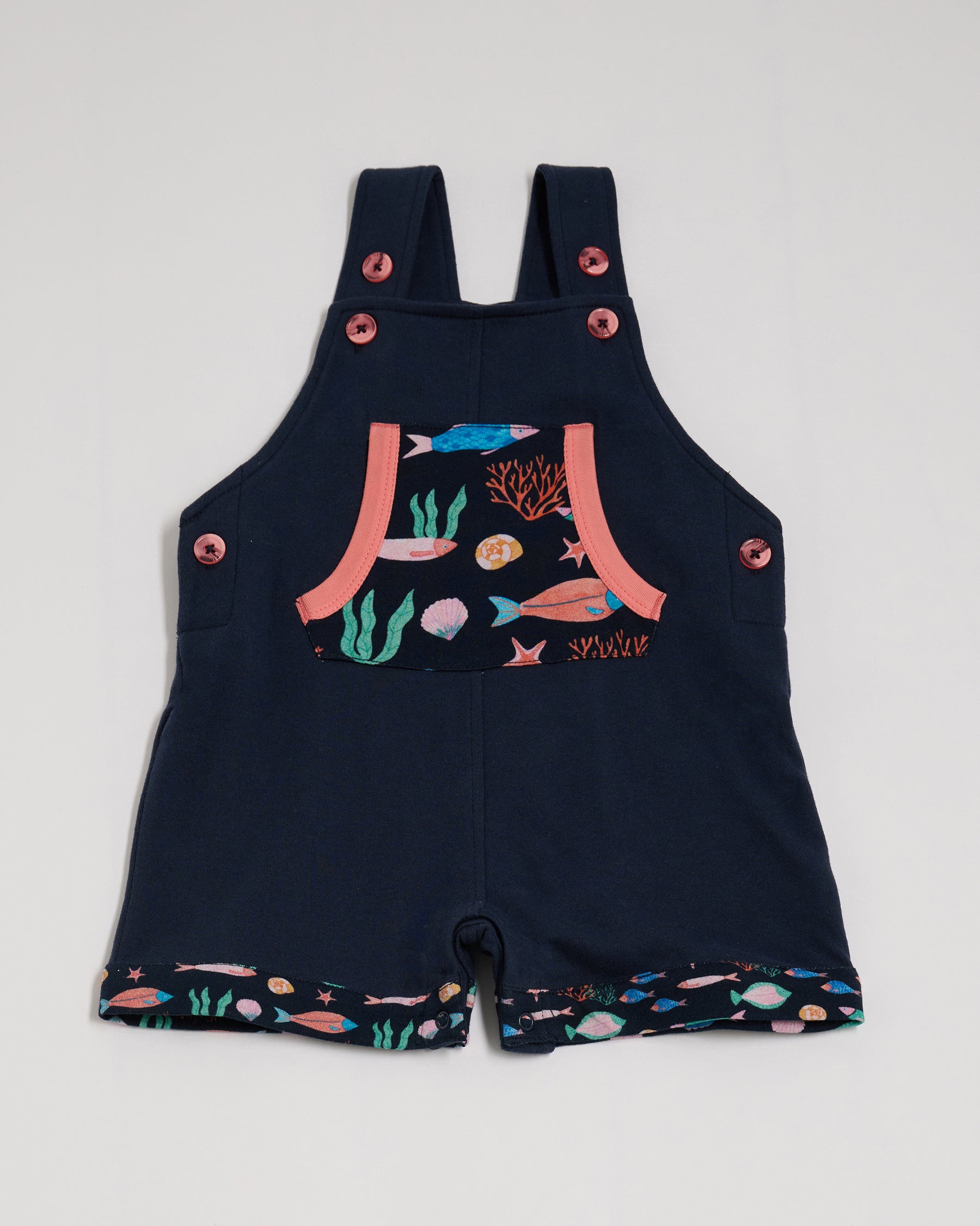 Front view of short dungarees with vibrant fish print pocket and fixed turn-ups, adjustable straps, crotch poppers, coral buttons and coral trim.