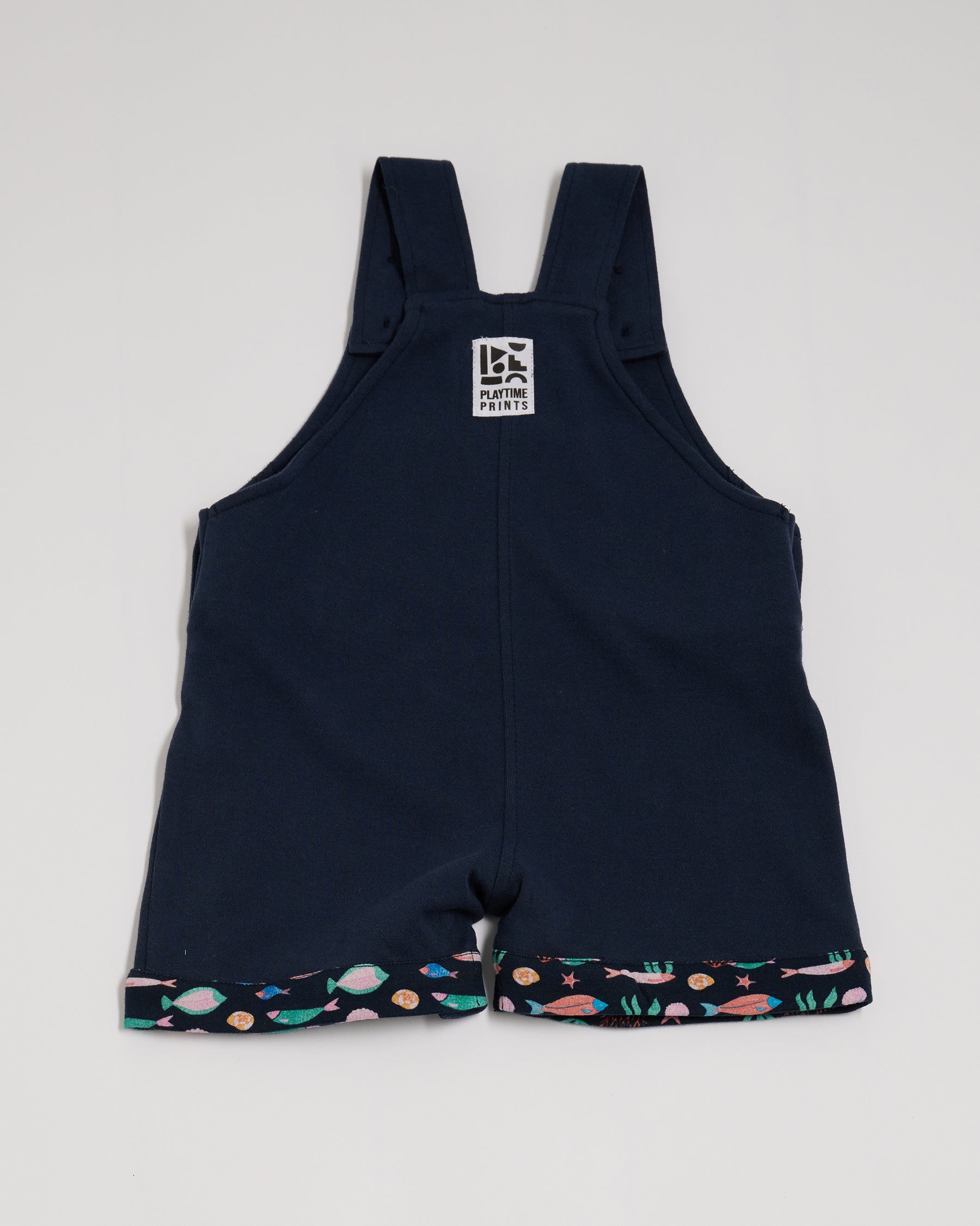 Back of navy short dungarees with fish print fixed turn-ups and Playtime Prints brand label.