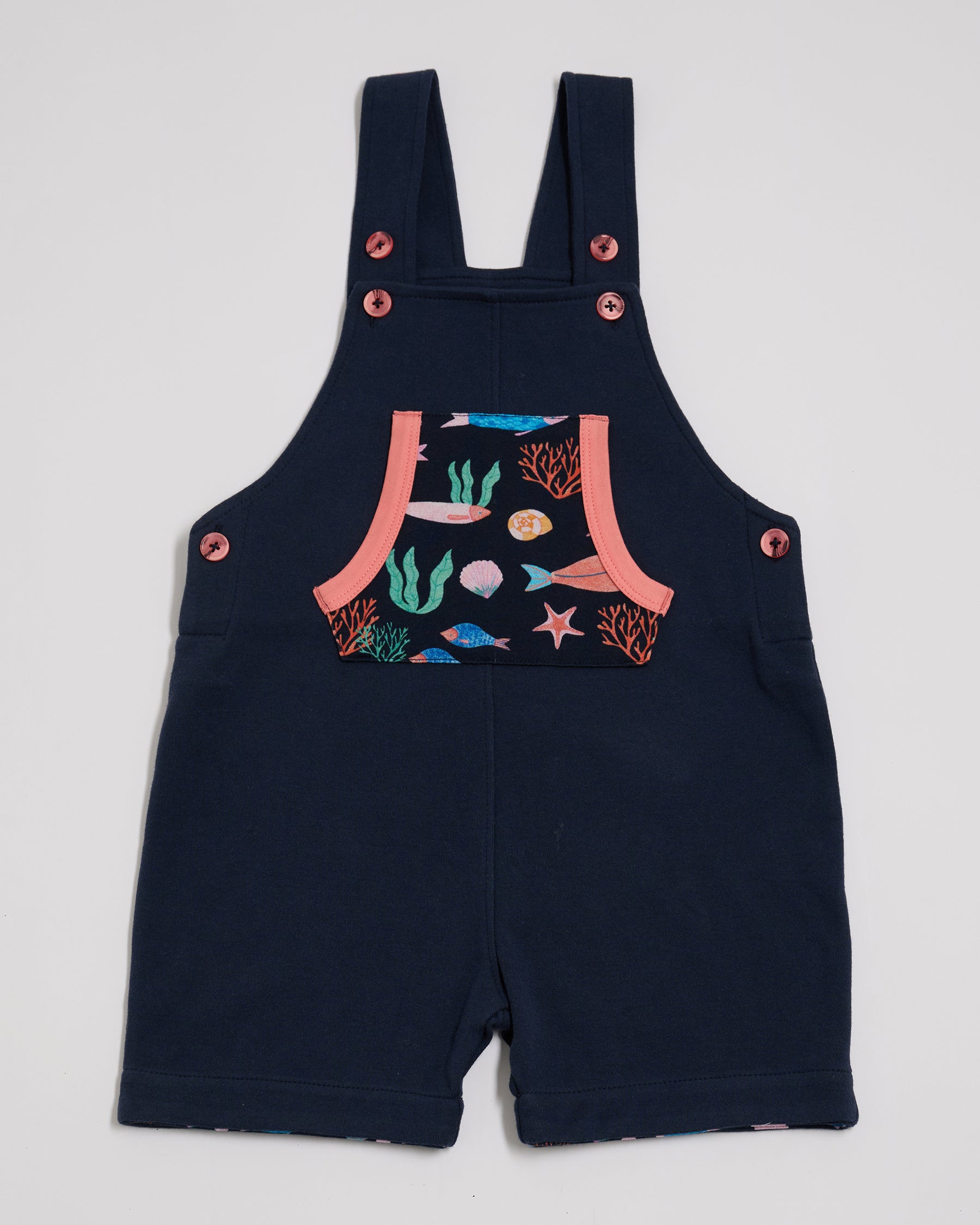 Front of navy short dungarees with vibrant fish print pocket, coral buttons and coral trim.