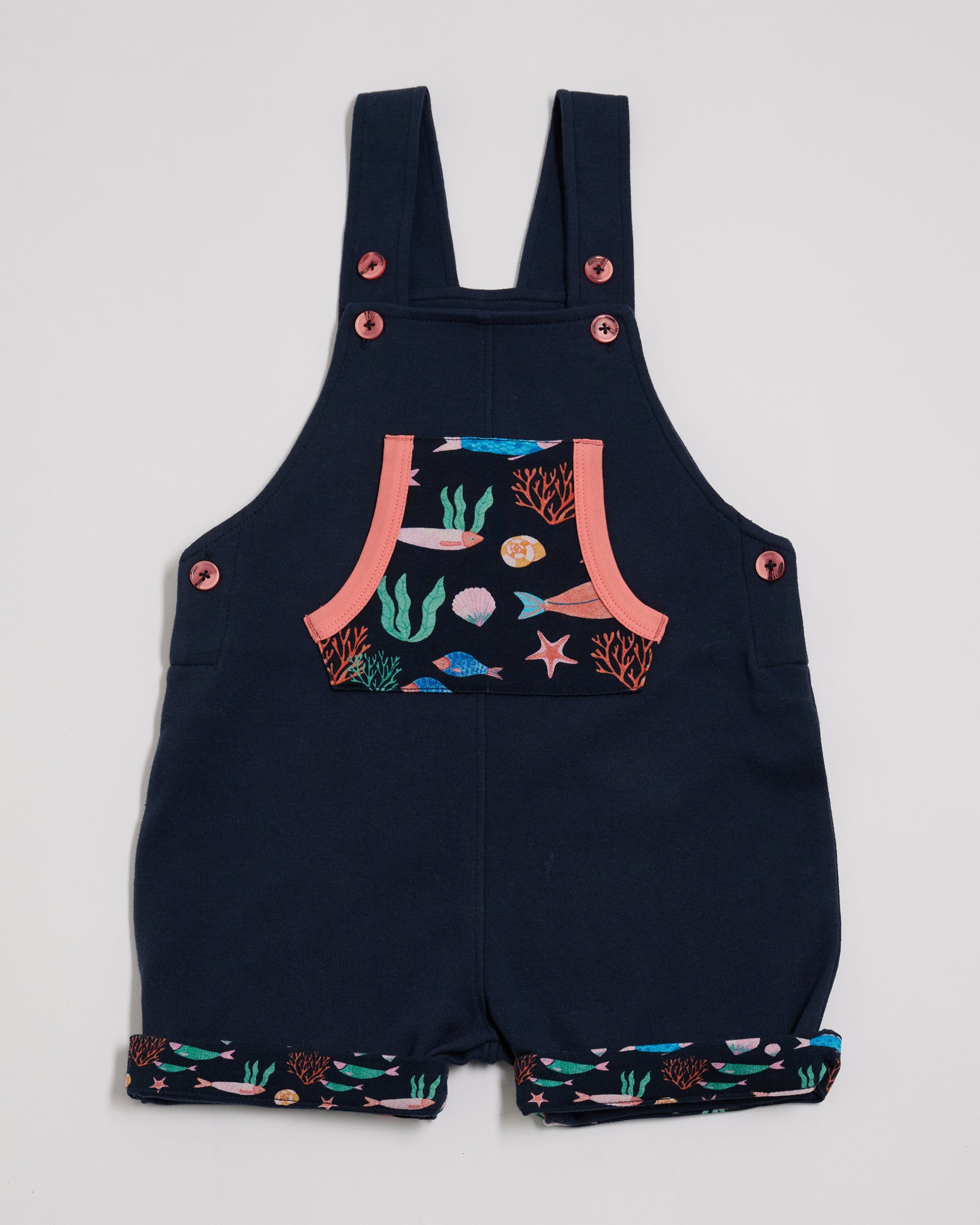 Front view of short dungarees with vibrant fish print pocket and turn-ups, adjustable height straps, coral buttons and coral trim.