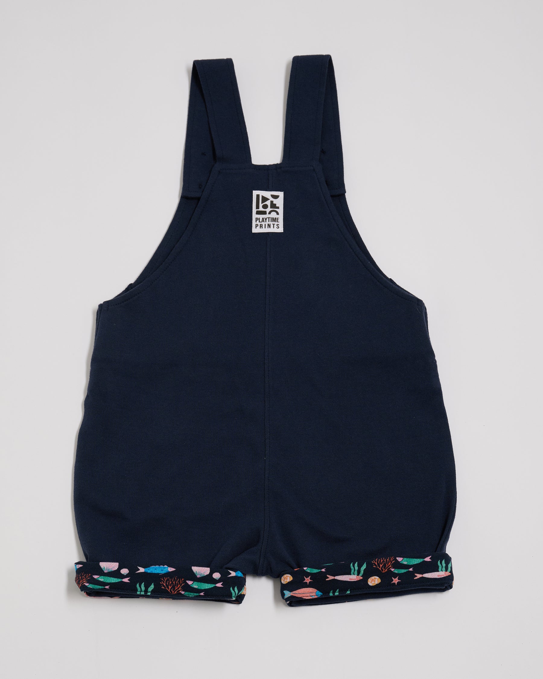 Back of navy short dungarees with fish print turn-ups and Playtime Prints brand label.