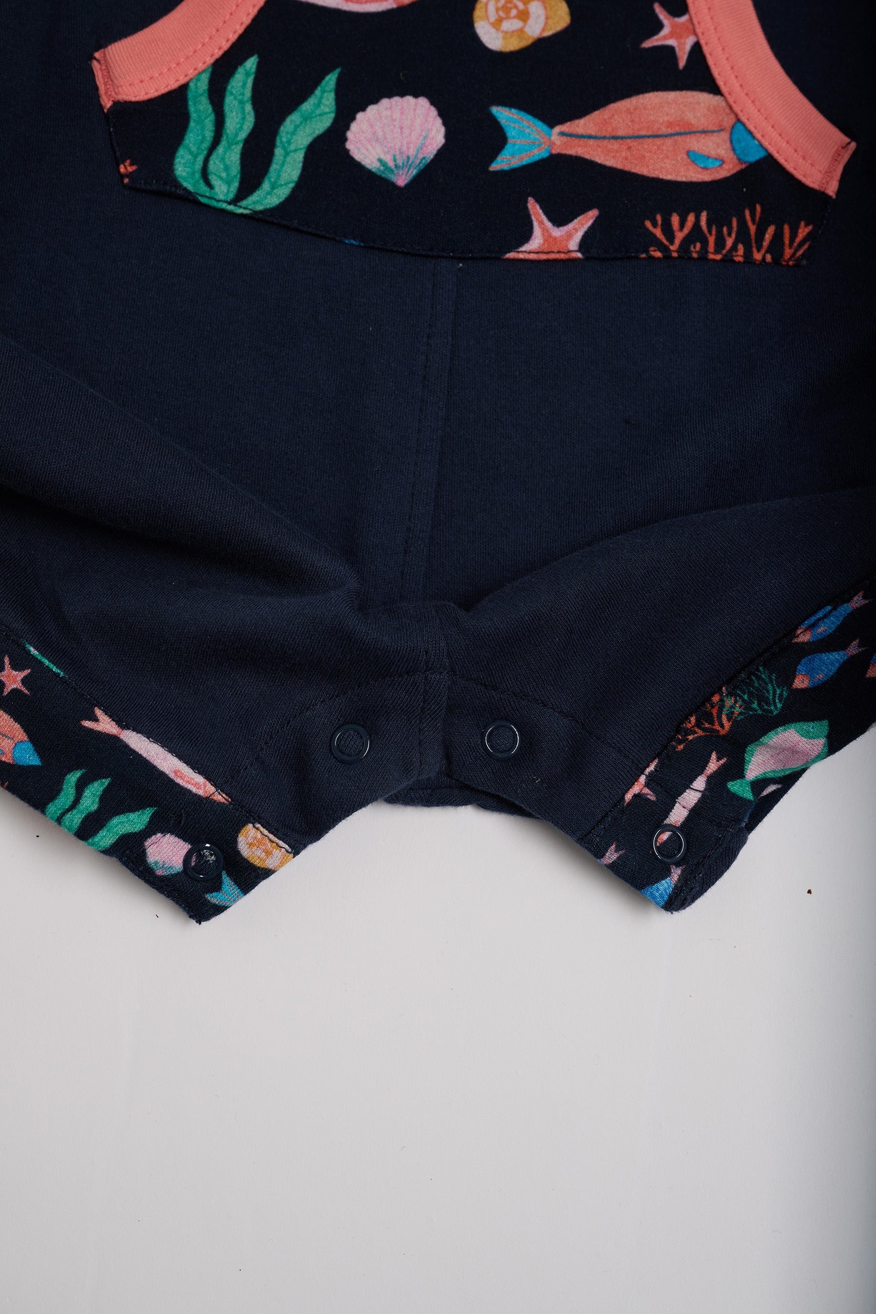 Close-up of crotch poppers on navy short dungarees with vibrant fish print pocket and turn-ups and a coral trim.
