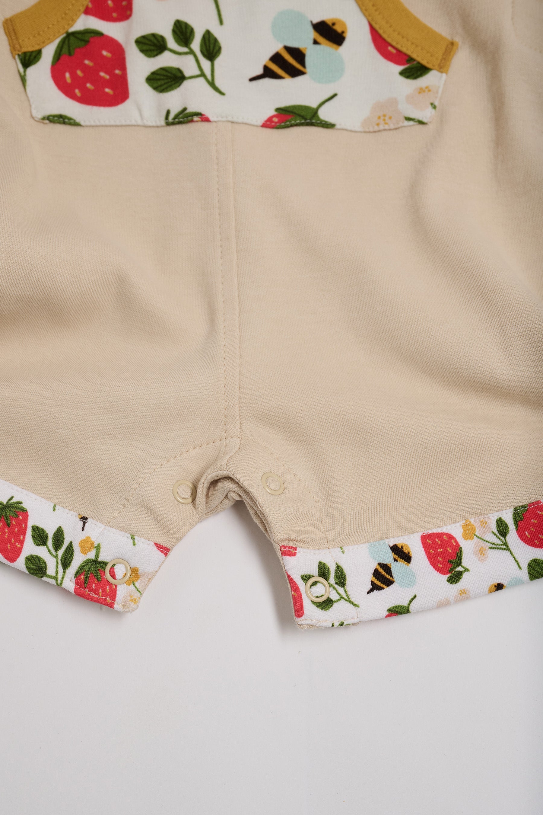 Close-up of crotch poppers on beige short dungarees with fun strawberry and bees print pocket and turn-ups and a honey trim.