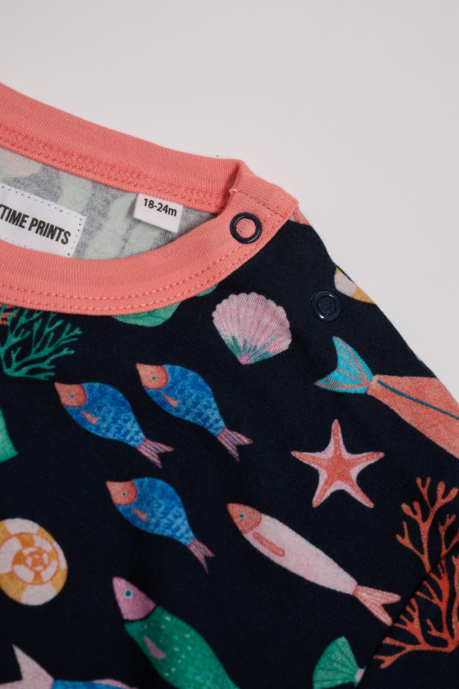 Close-up of neck poppers on a short sleeved t-shirt featuring an all over vibrant navy fish print with contrast coral neck and sleeve trim.