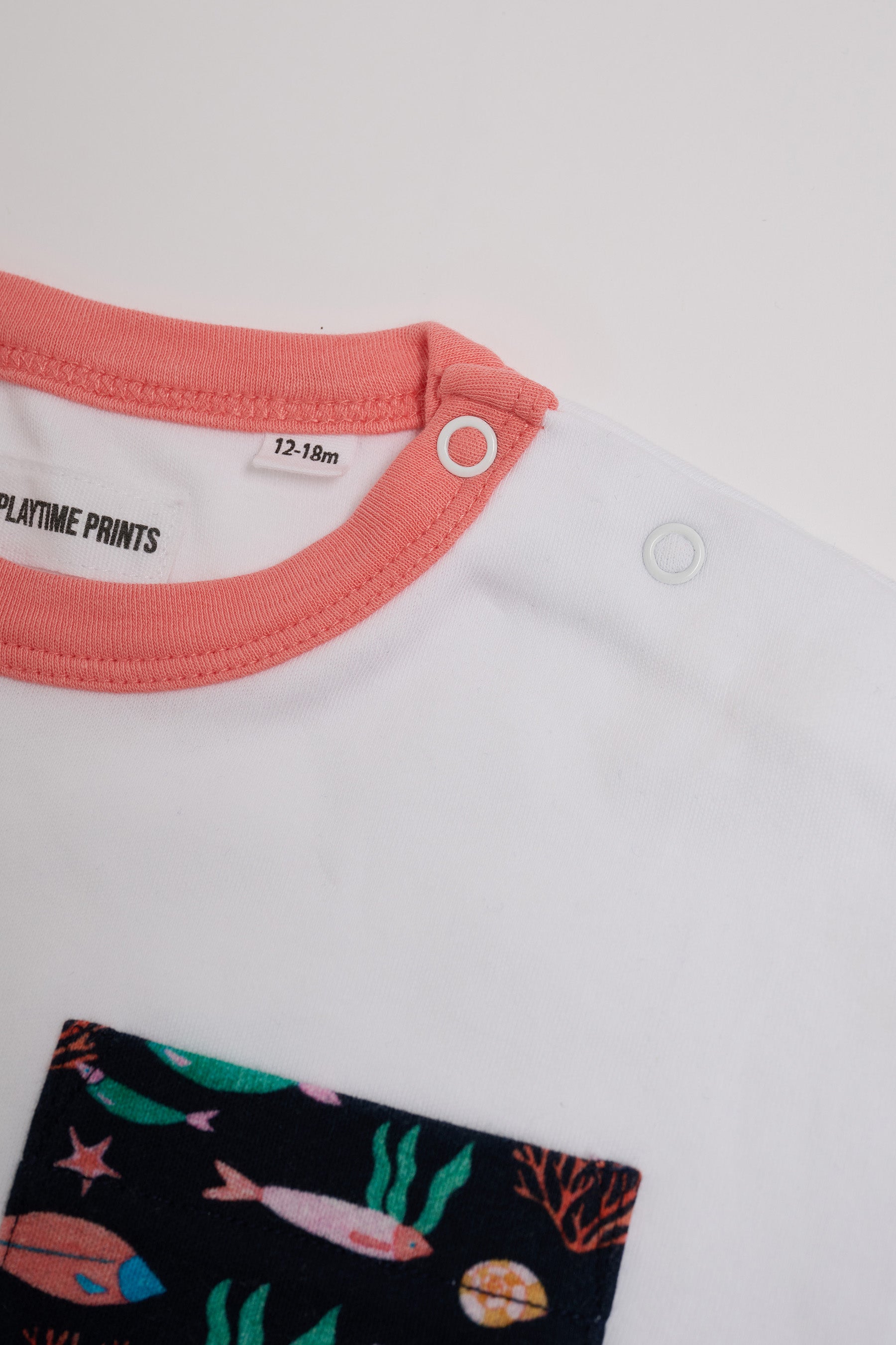 Close-up of neck poppers on white t-shirt with vibrant fish print chest pocket and coral neck trim.