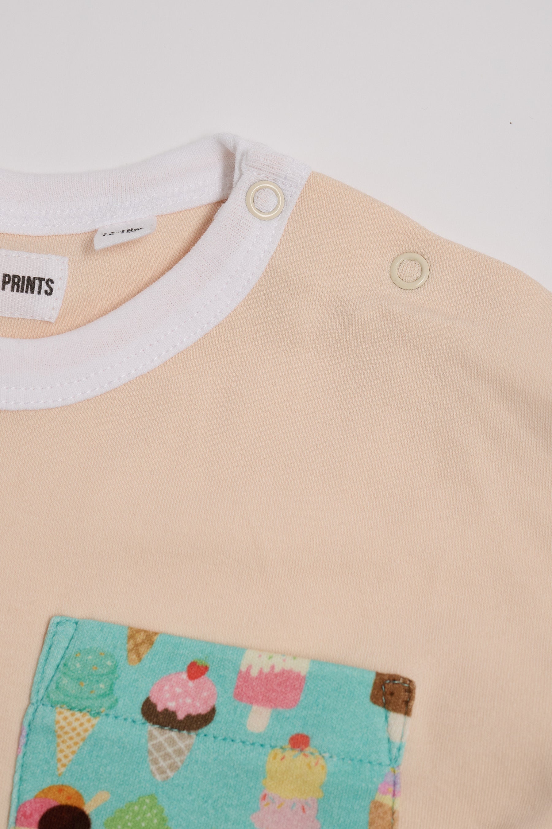 Close-up of neck poppers on vanilla coloured t-shirt with bright ice cream print square chest pocket and white neck trim.