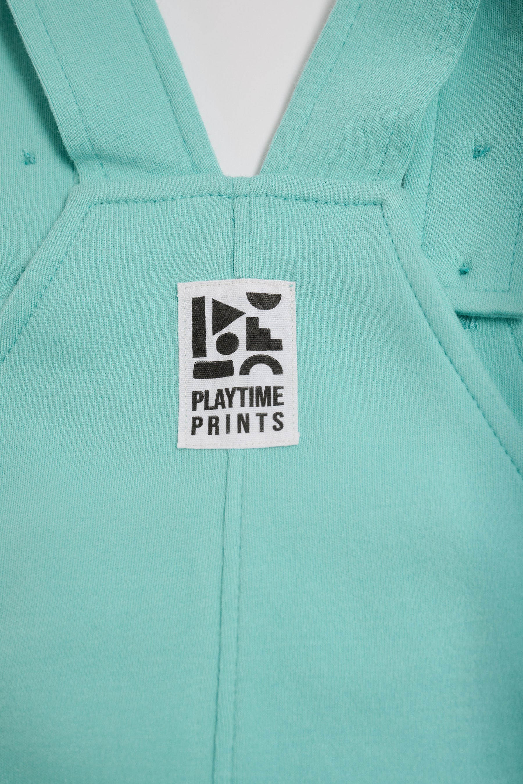 Close-up of Playtime Prints brand label on the back of turquoise short dungarees.