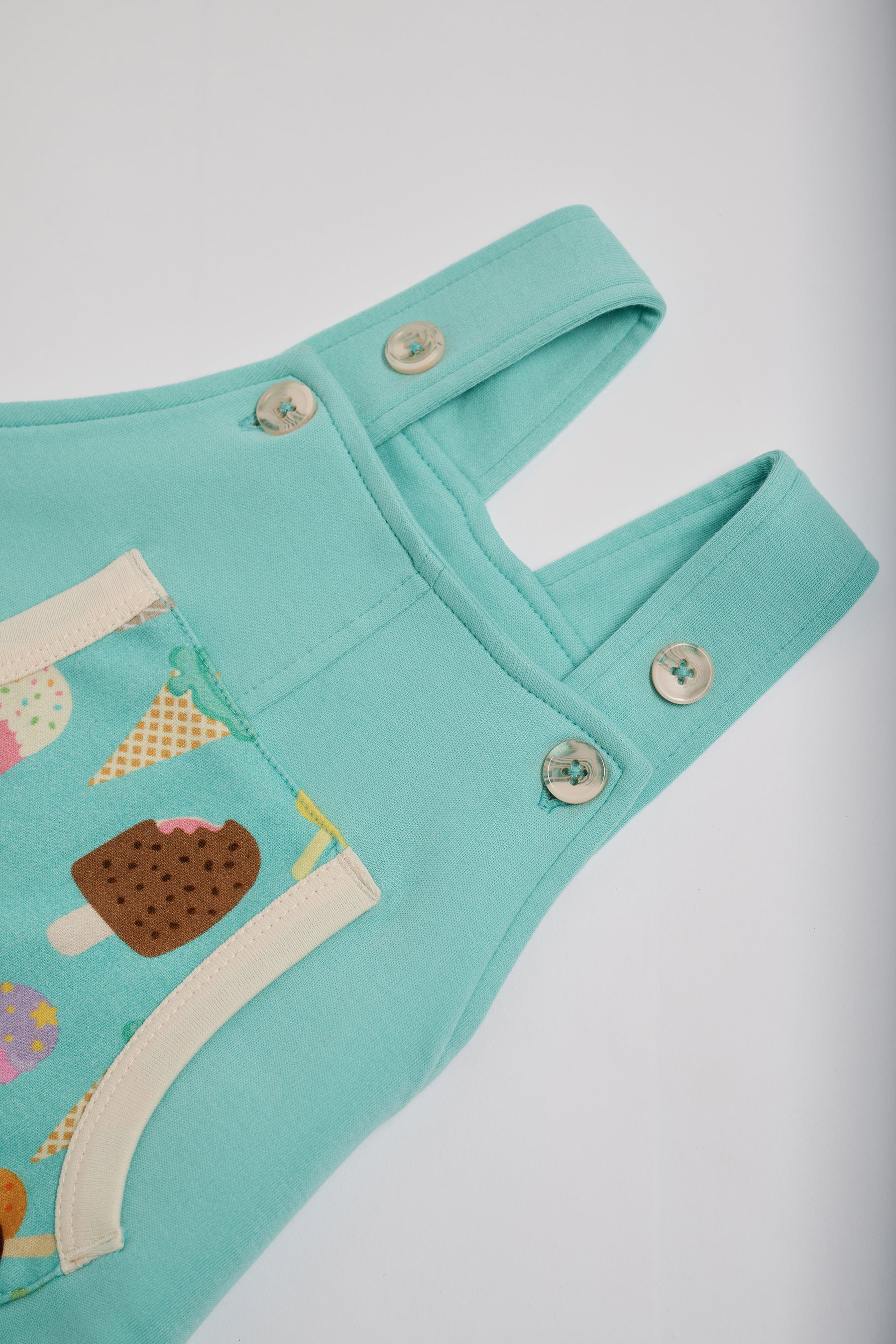High setting on adjustable straps on turquoise short dungarees with bright ice cream print pocket, vanilla buttons and vanilla trim.