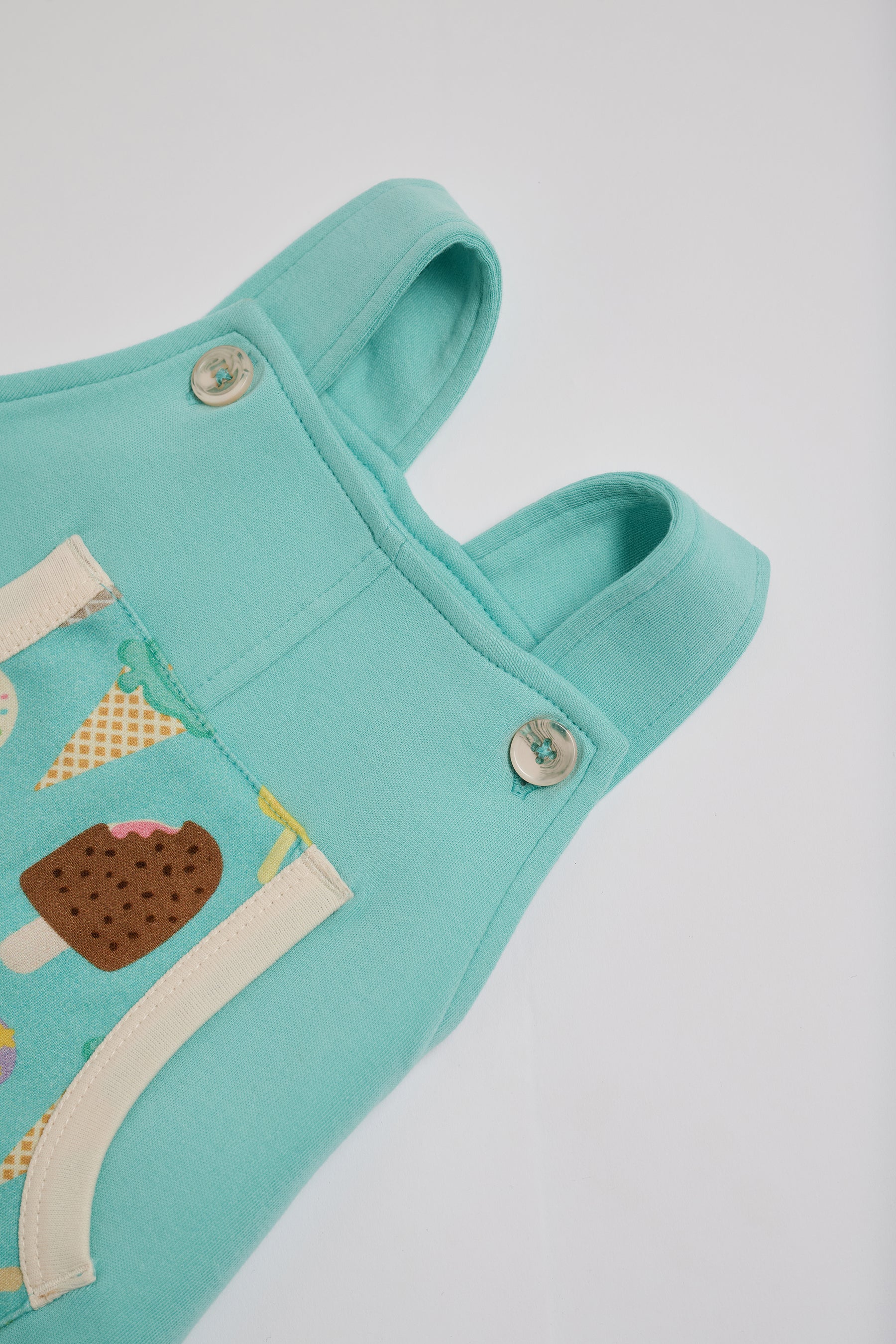 High setting on adjustable straps on turquoise short dungarees with bright ice cream print pocket, vanilla buttons and vanilla trim.