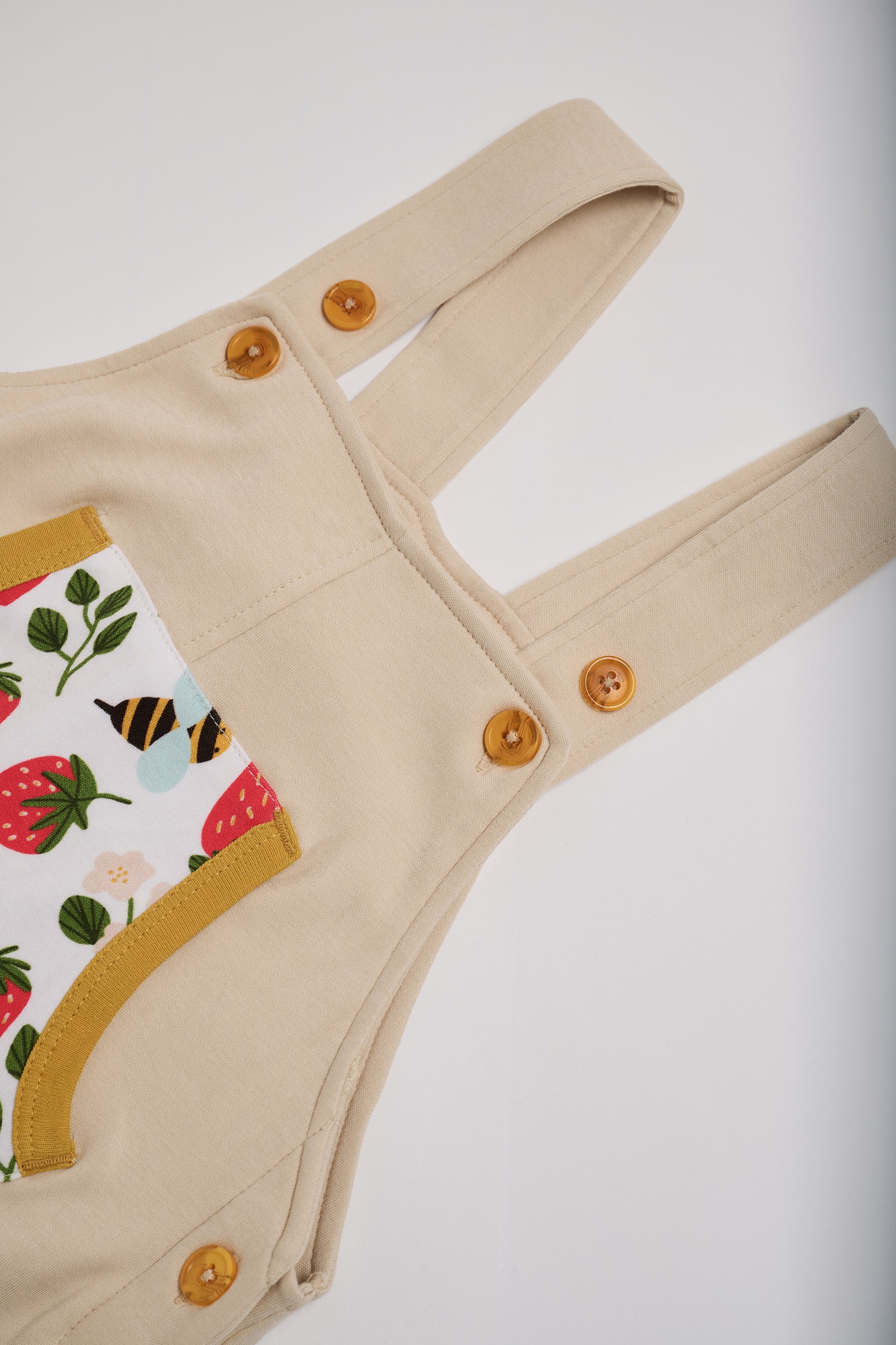 Close-up of adjustable straps set to high setting on beige short dungarees with fun strawberry and bees print and honey buttons and honey trim.