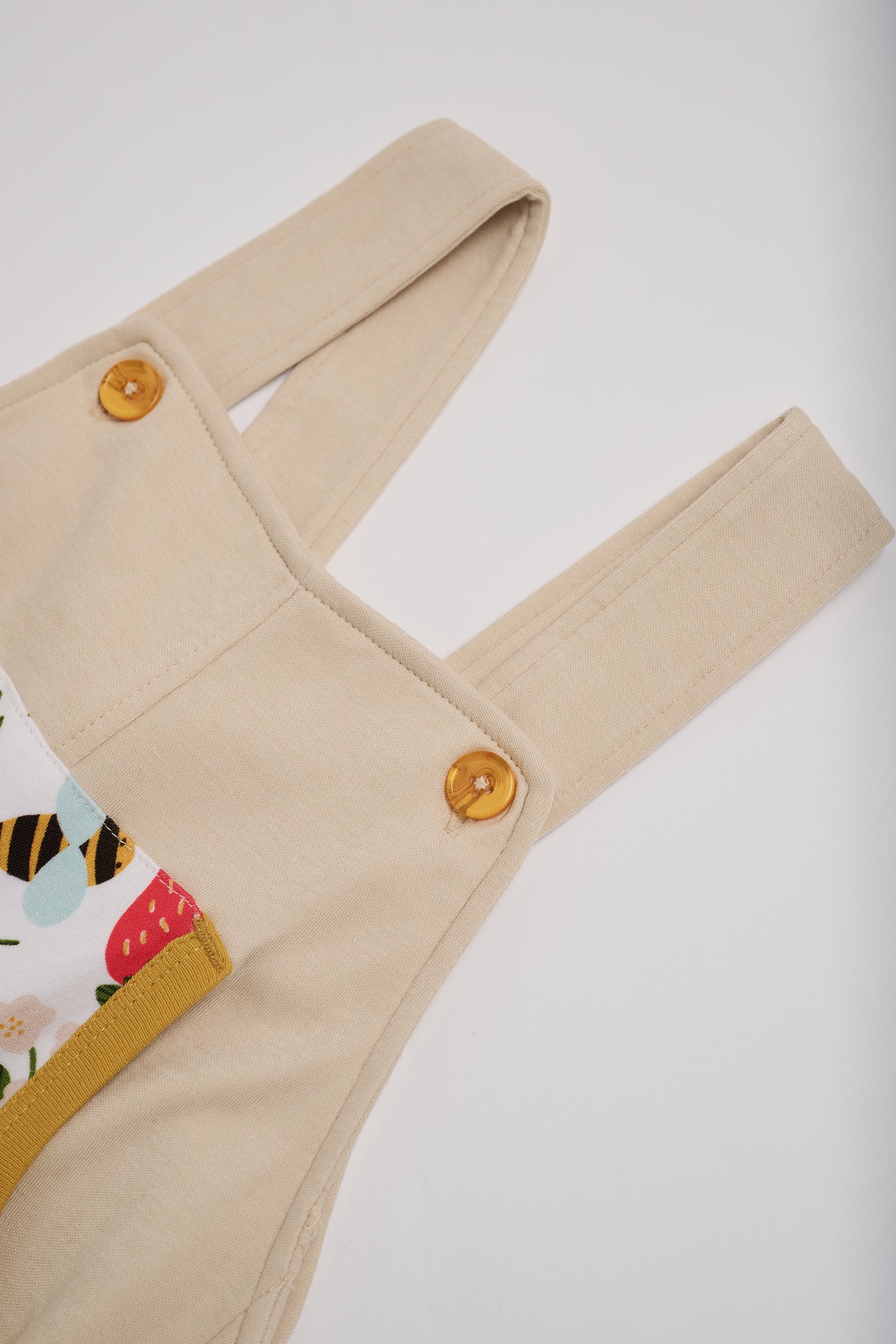 Close-up of adjustable straps set to short setting on beige short dungarees with fun strawberry and bees print and honey buttons and honey trim.