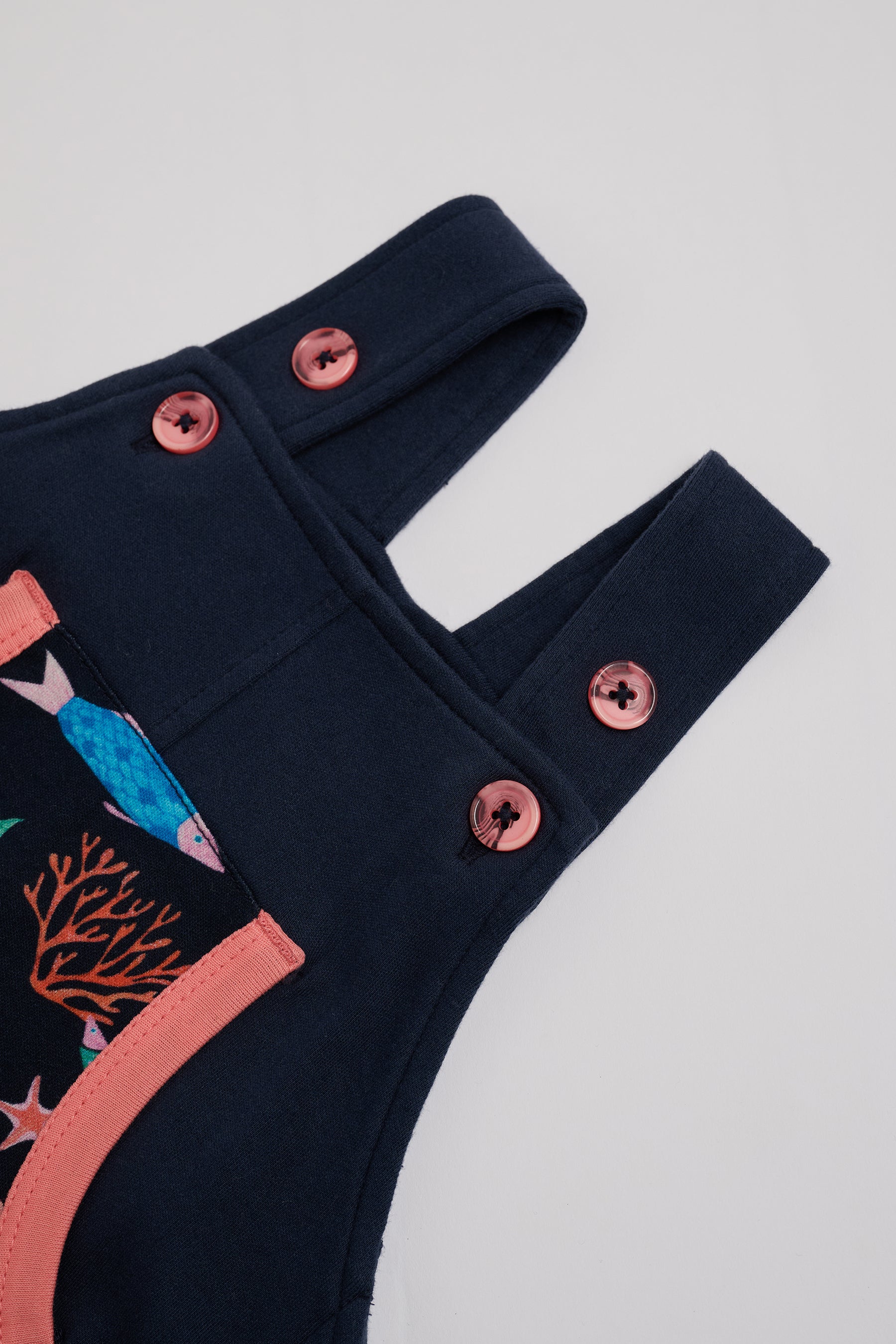 Close-up of adjustable straps set to high setting on navy short dungarees with vibrant fish print and coral buttons and coral trim.
