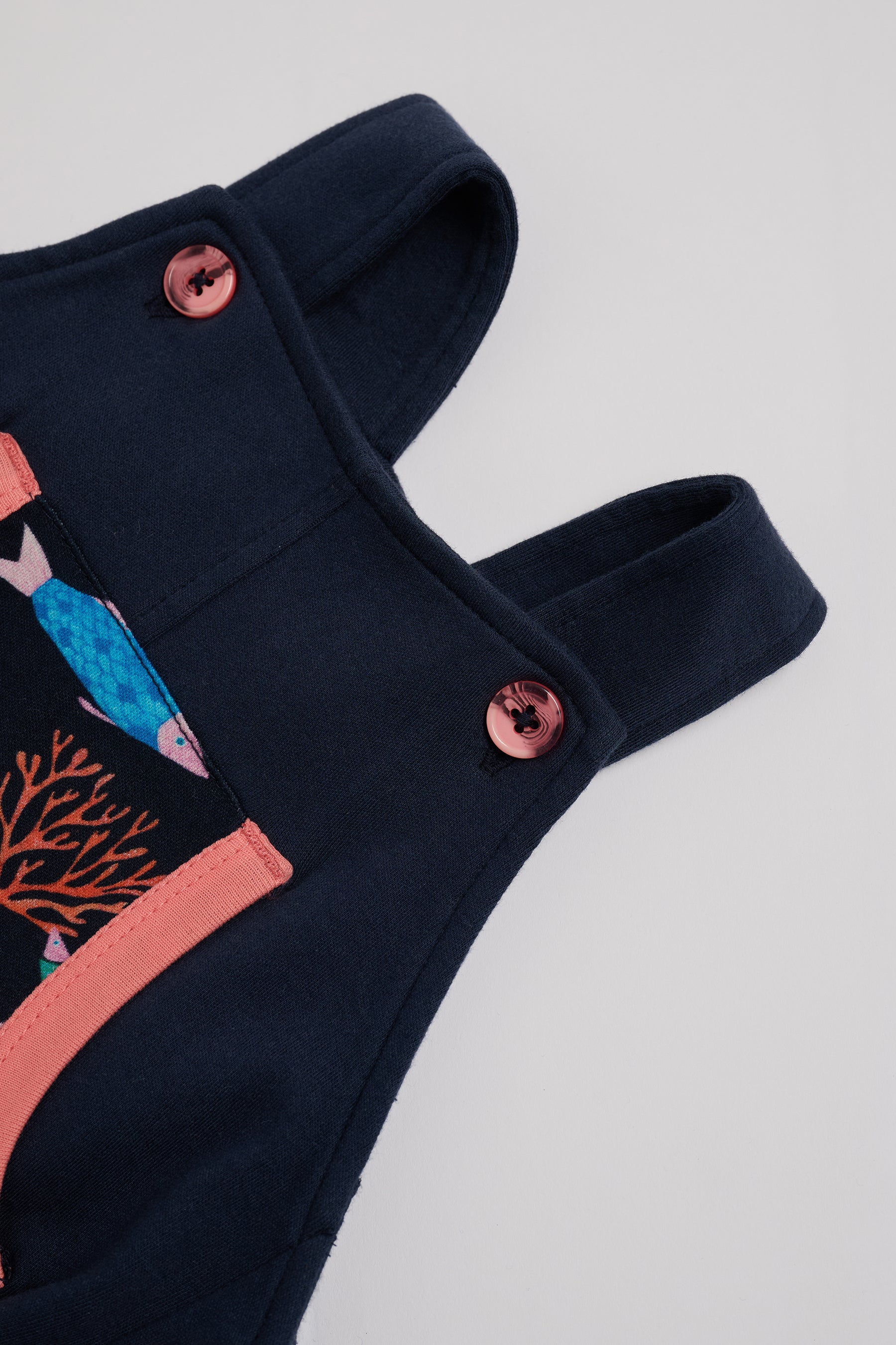 Close-up of adjustable straps set to short setting on navy short dungarees with vibrant fish print and coral buttons and coral trim.