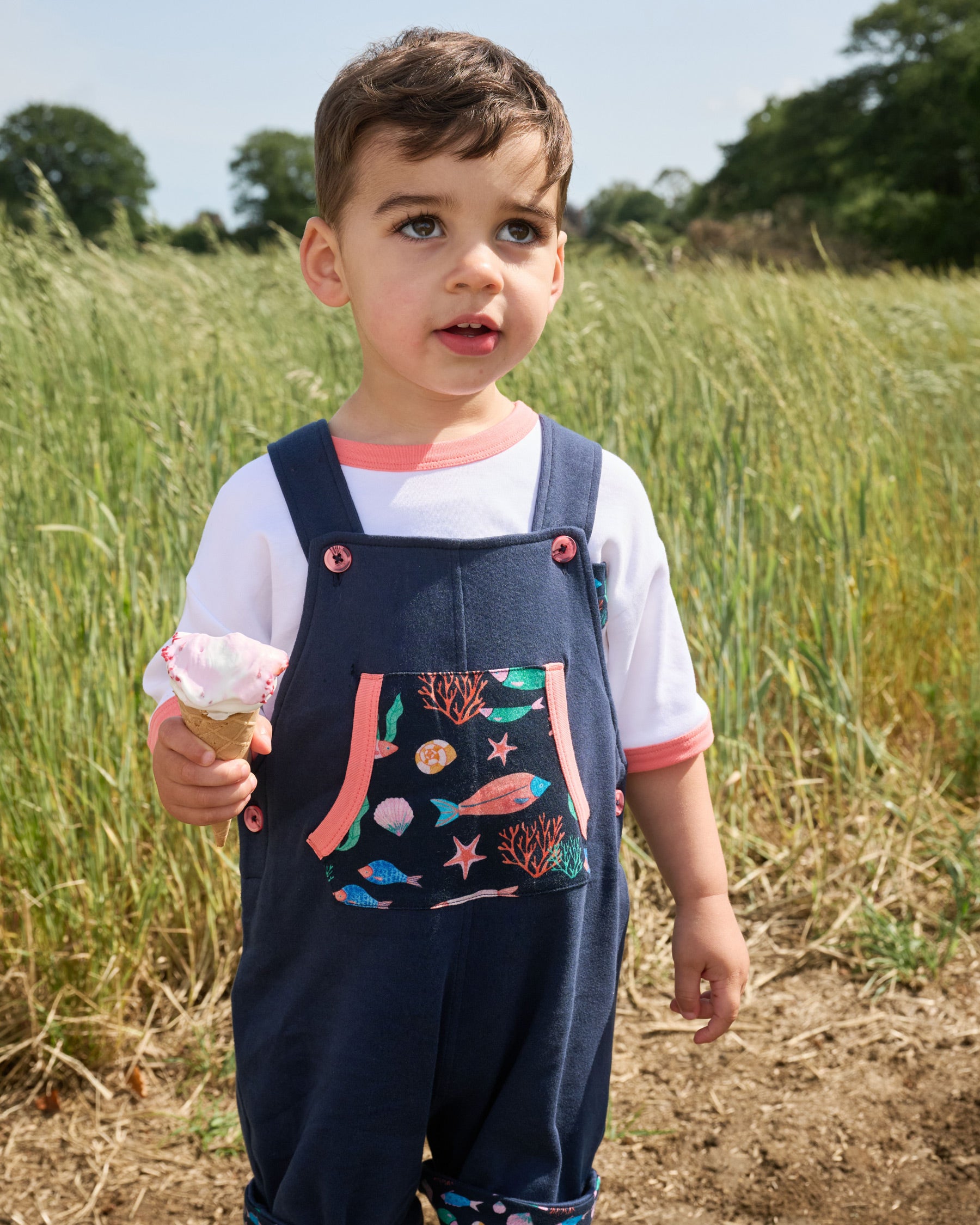 Young boy wearing navy short dungarees with vibrant fish print pocket, coral buttons and coral trim along with a white t-shirt with coral trim.