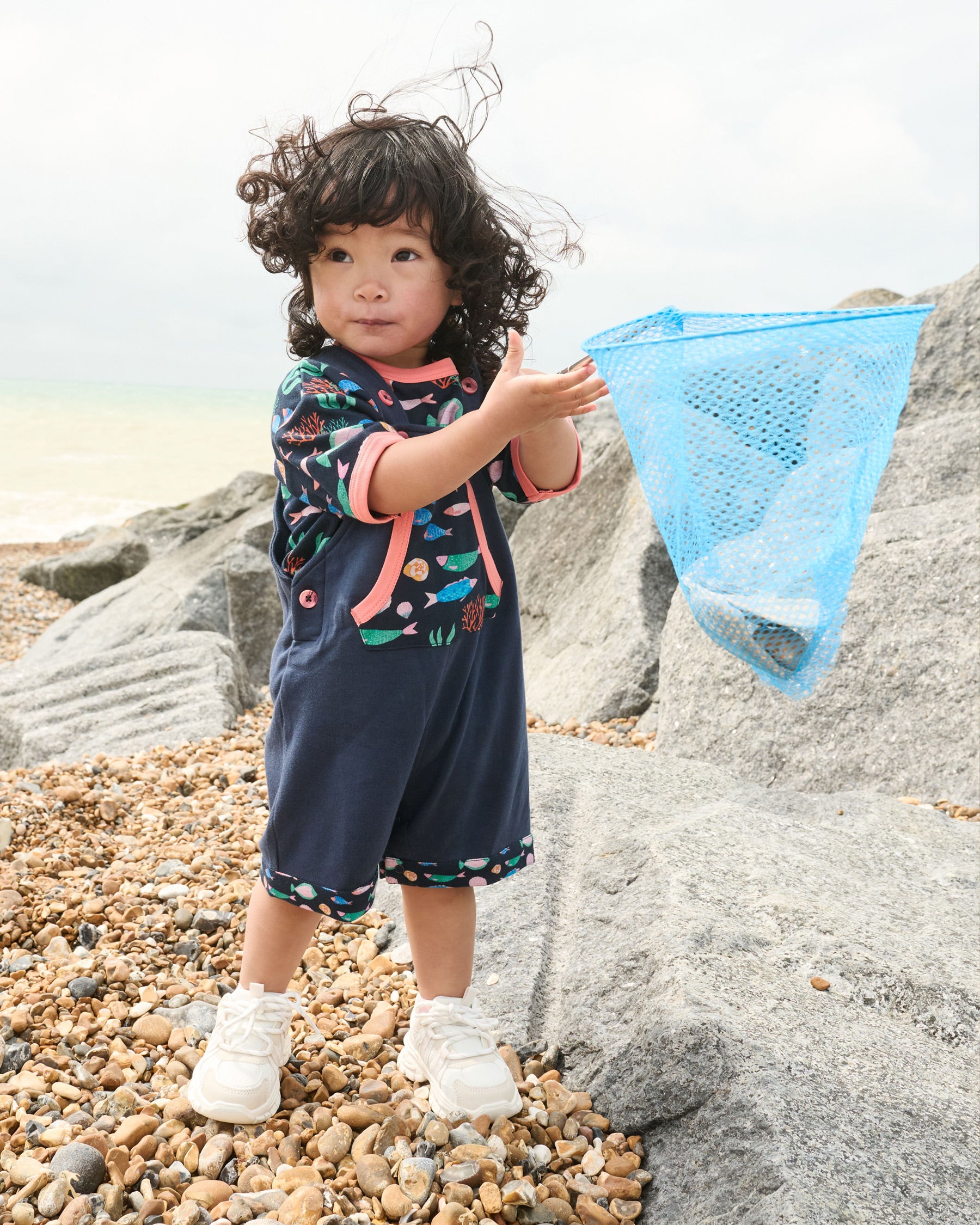 Little girl holding a fishing net and wearing a vibrant navy fish print short sleeved t-shirt with contrast coral neck and sleeve trim under a set of matching navy dungarees.