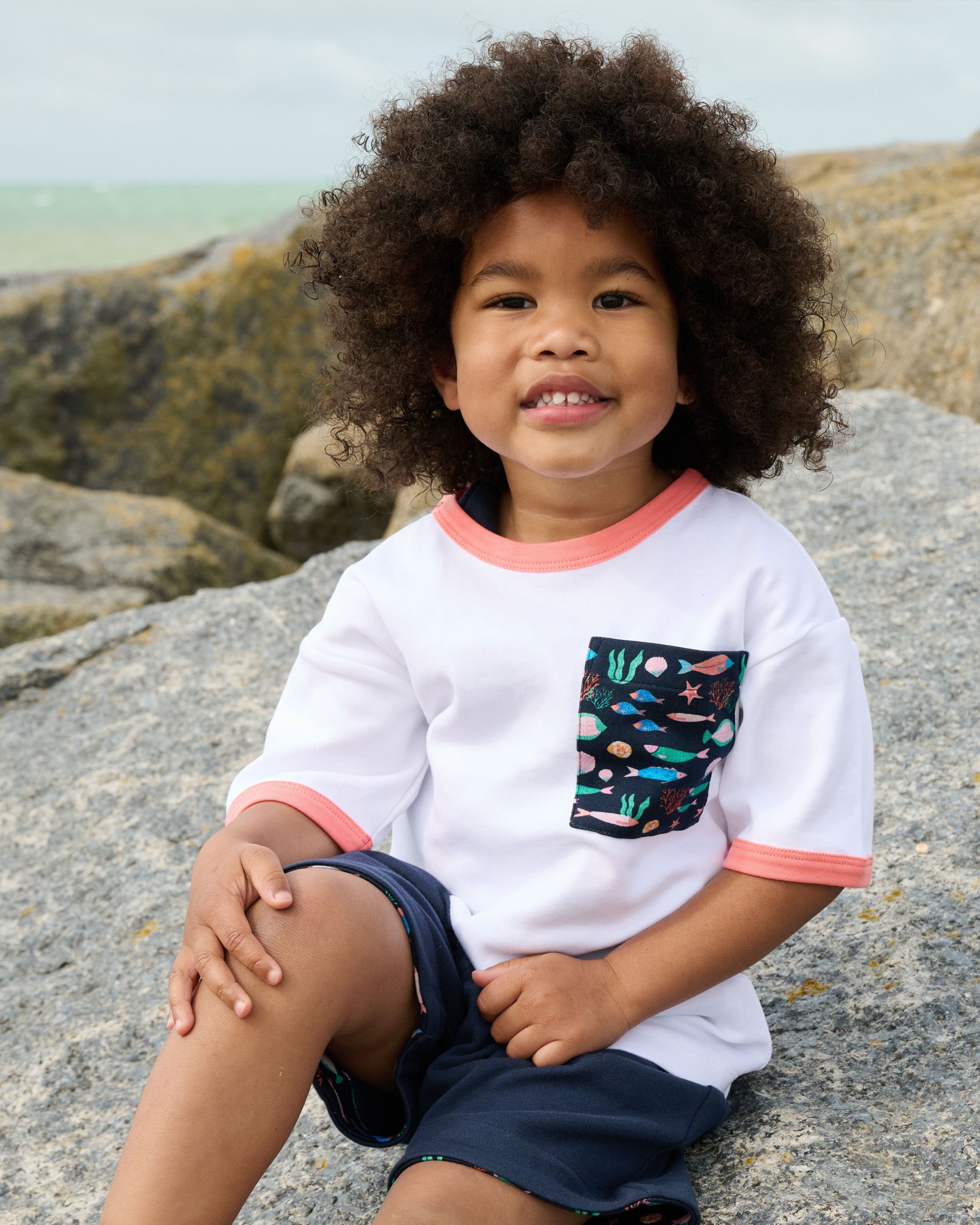Young boy smiling wearing a white short sleeved t-shirt with vibrant fish print square chest pocket and coral neck and sleeve trim over navy short dungarees.