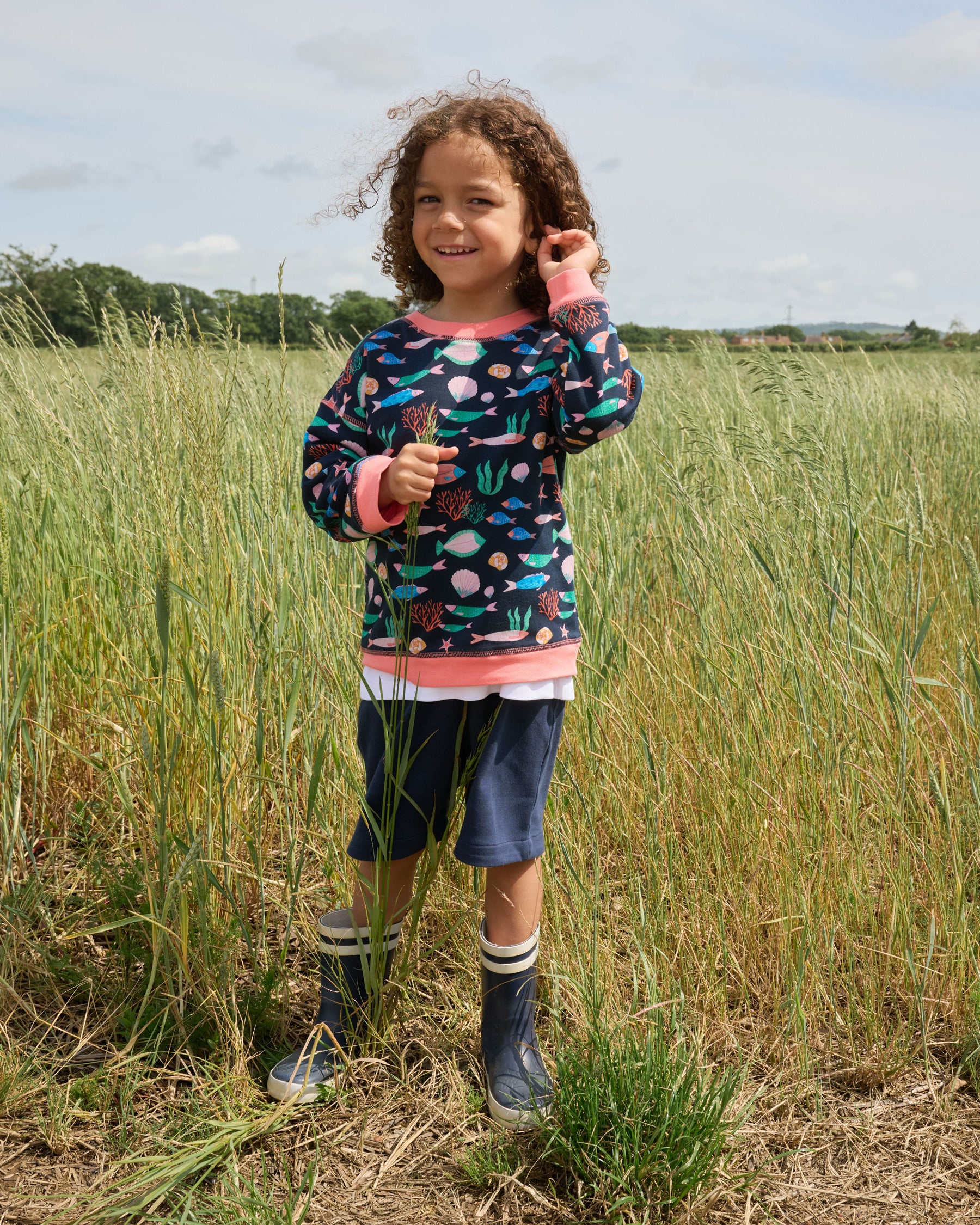 Young boy in field smiling at the camera and wearing a sweatshirt featuring an all over vibrant navy fish print with contrast coral neck and sleeve trim over a set of matching navy short dungarees.