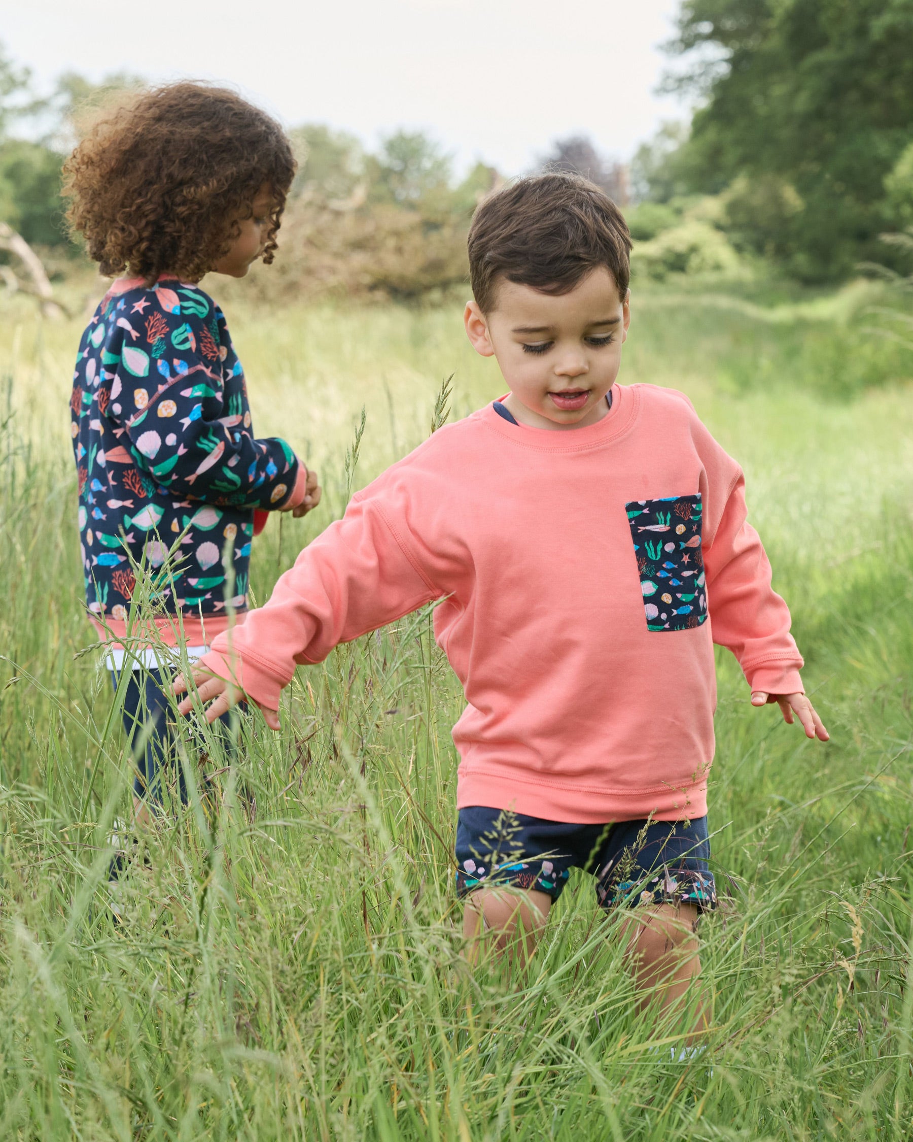 Two boys playing in a field - one wearing a sweatshirt featuring an all over vibrant navy fish print with contrast coral neck and sleeve trim and the other a coral sweatshirt with an oversized vibrant navy fish print chest pocket.