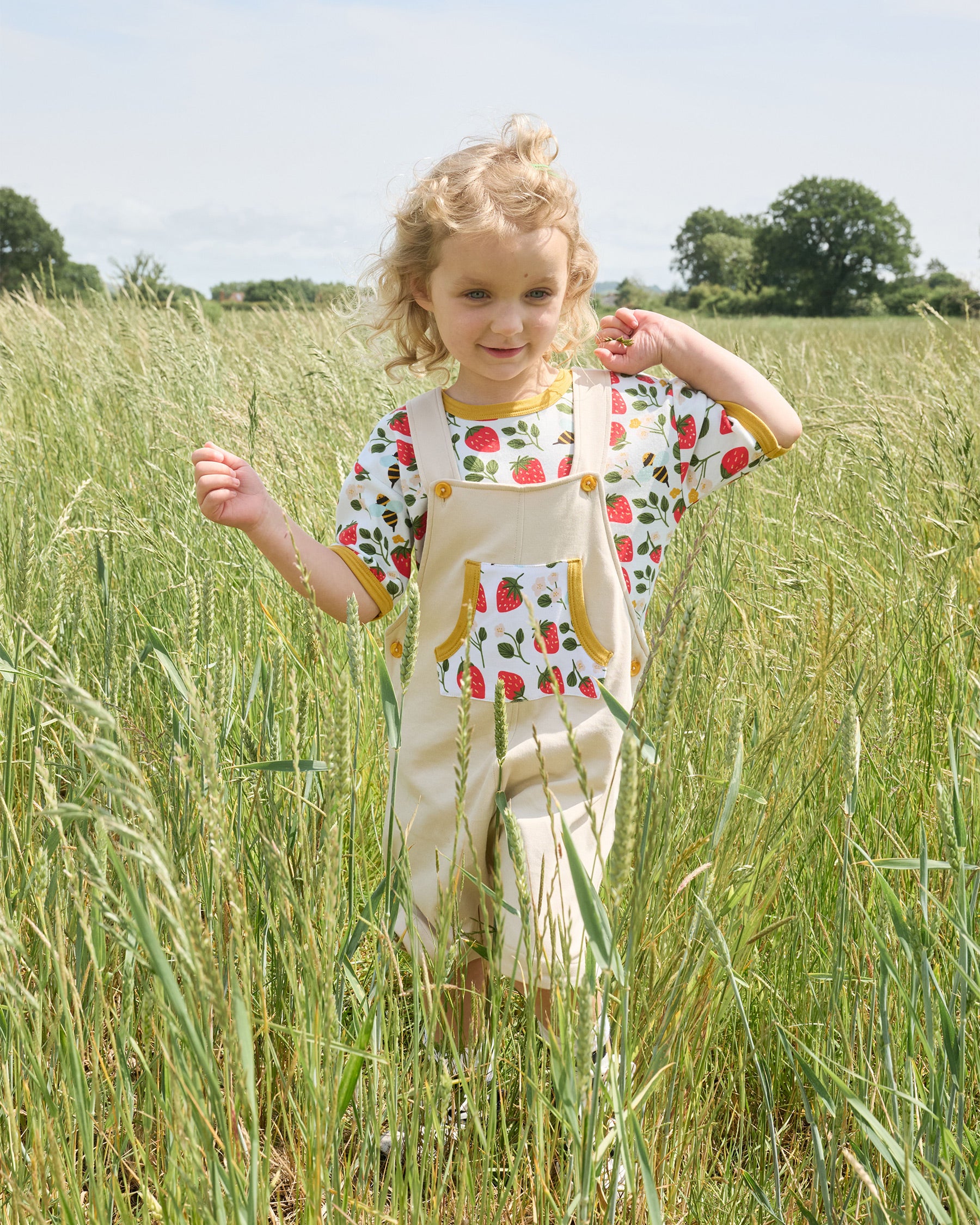 Young girl in a field wearing beige short dungarees with a fun strawberry and bee pocket, honey buttons and honey trim. Underneath the dungarees she wears a full print strawberry and bees t-shirt.