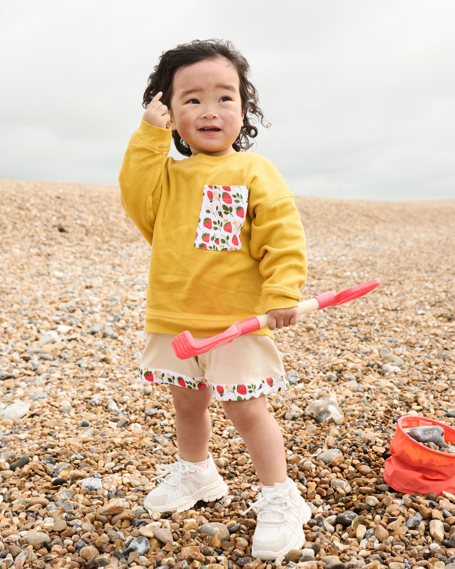 Little girl on the beach wearing a honey coloured sweatshirt with an oversized strawberry and bees print chest pocket over a set of matching beige dungarees.