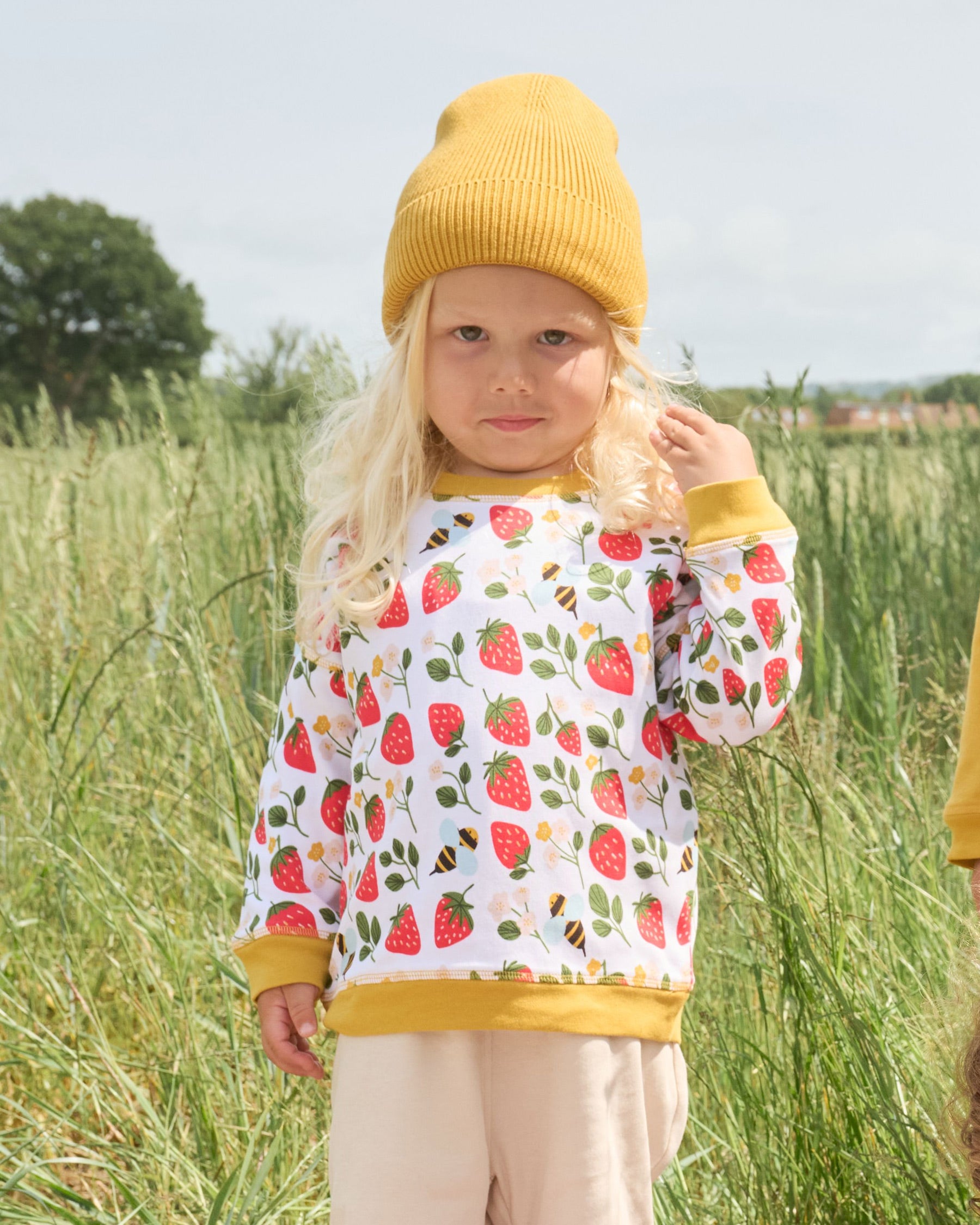 Little boy in a field wearing a honey coloured hat and a sweatshirt featuring an all over strawberry and bees print with contrast honey neck and sleeve trim over a set of matching beige dungarees.